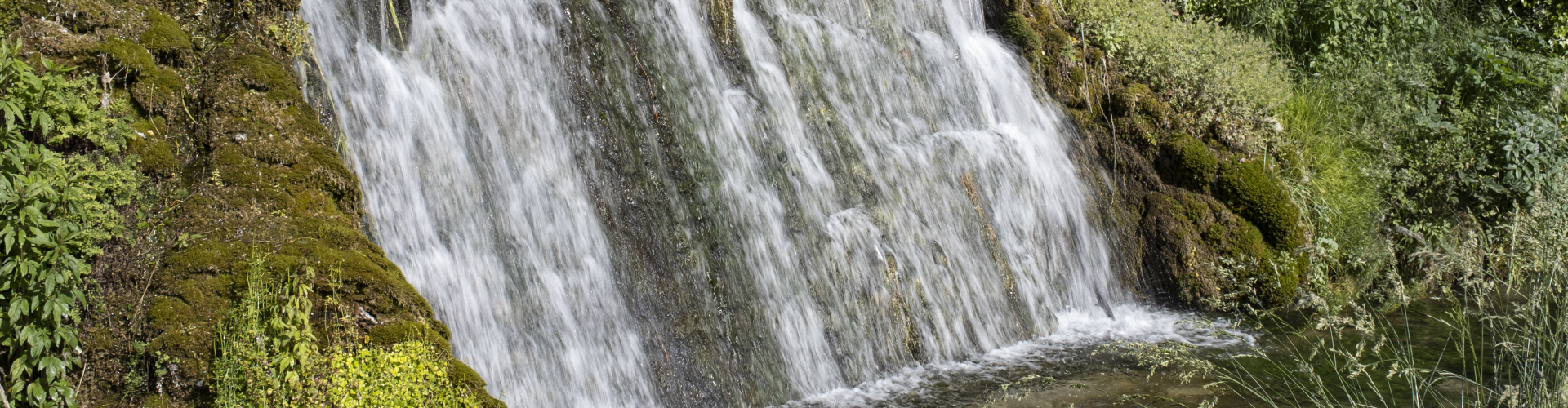 Cascata che scende vigorosamente tra pareti rocciose, formando un piccolo bacino d'acqua circondato da vegetazione. L'acqua spumeggiante crea un forte contrasto con le rocce ruvide e la fitta vegetazione della gola.