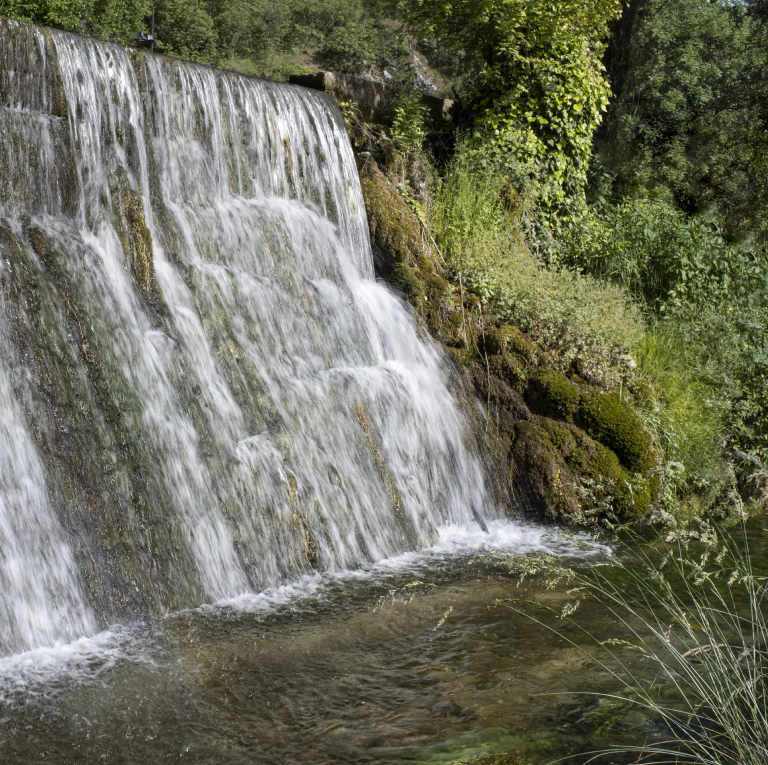 Cascata che scende vigorosamente tra pareti rocciose, formando un piccolo bacino d'acqua circondato da vegetazione. L'acqua spumeggiante crea un forte contrasto con le rocce ruvide e la fitta vegetazione della gola.